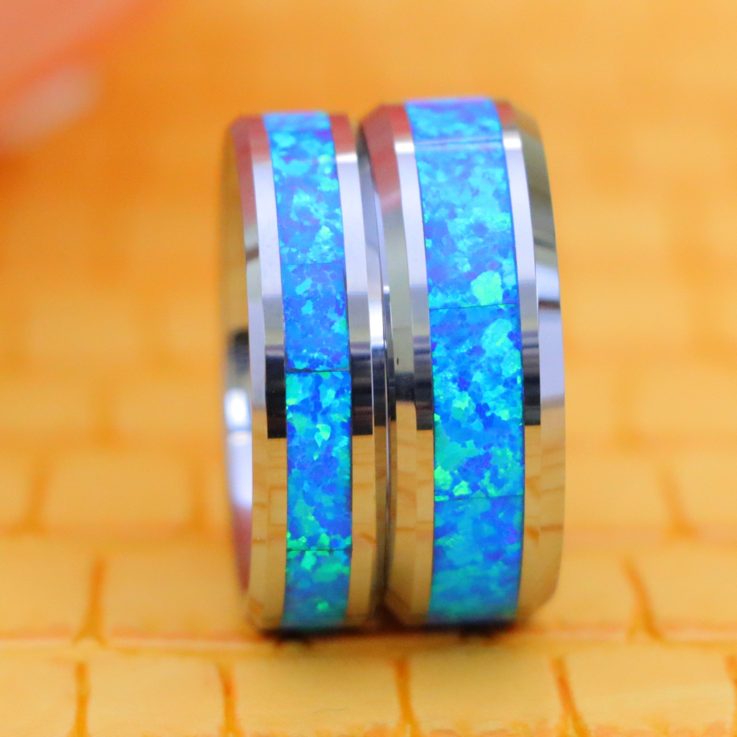 Two tungsten wedding bands with vibrant blue opal inlays set against a bright orange background - Men's Jewelry